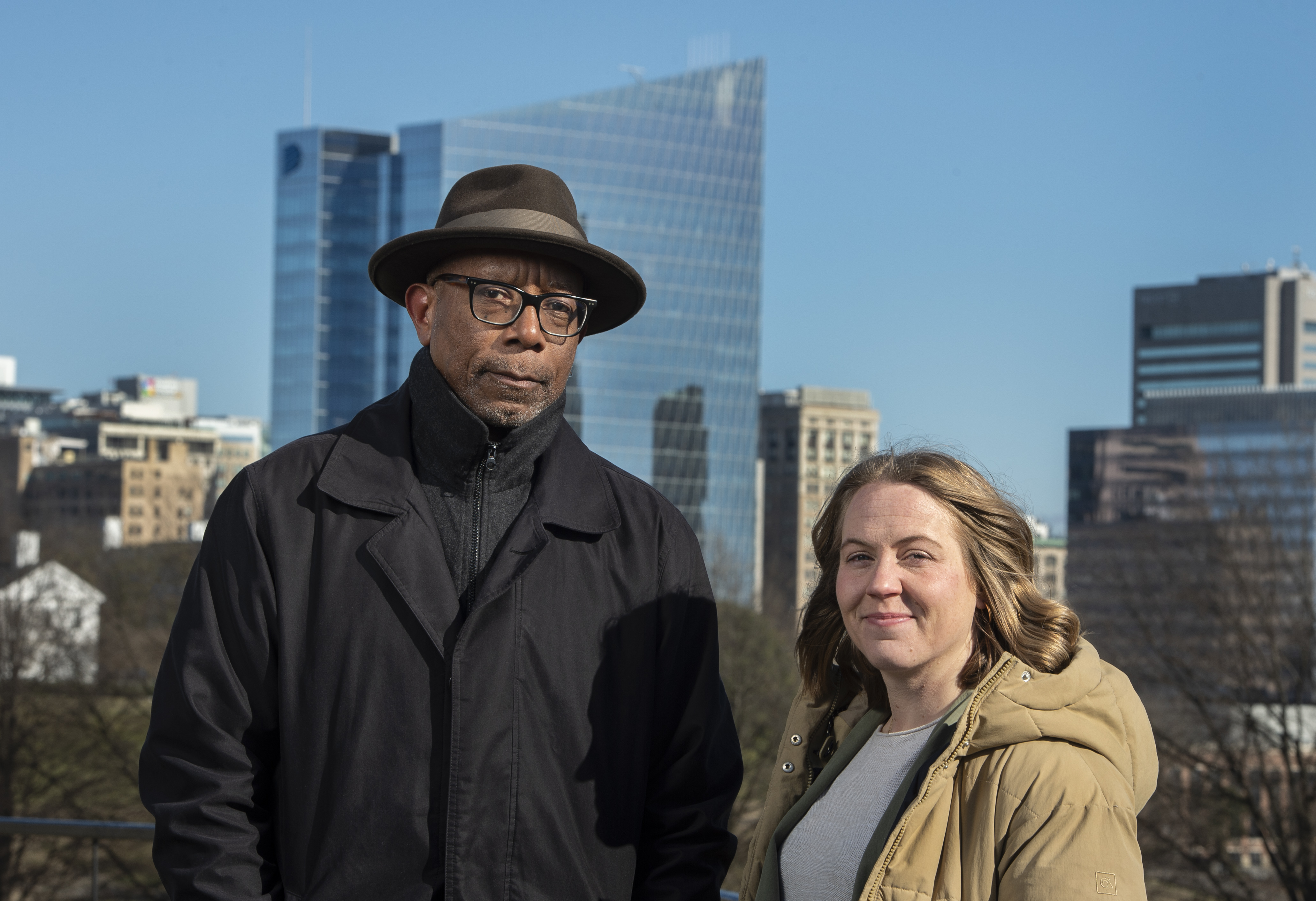 A man wearing a black pea coat and a rimmed hat stadning beside a woman in a light brown back and gray shirt. The Richmond skyline is in the background.