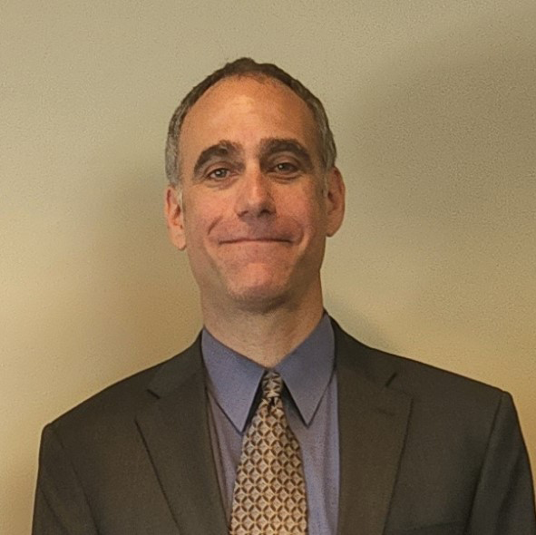 A man wearing a brow suit jacket, blue shirt and gold tie