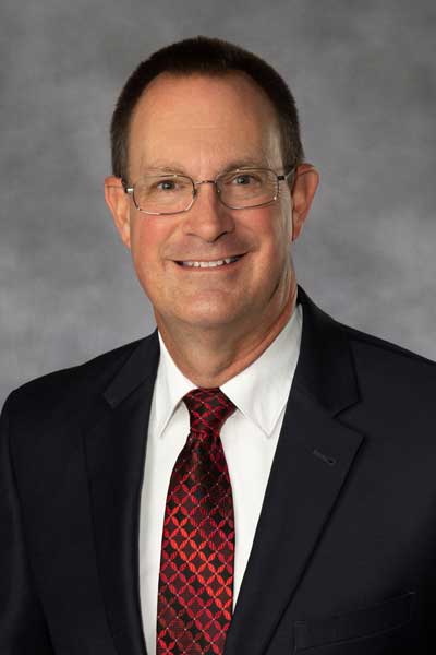 A man weating a black jacket, white dress shirt and red tie with glasses.