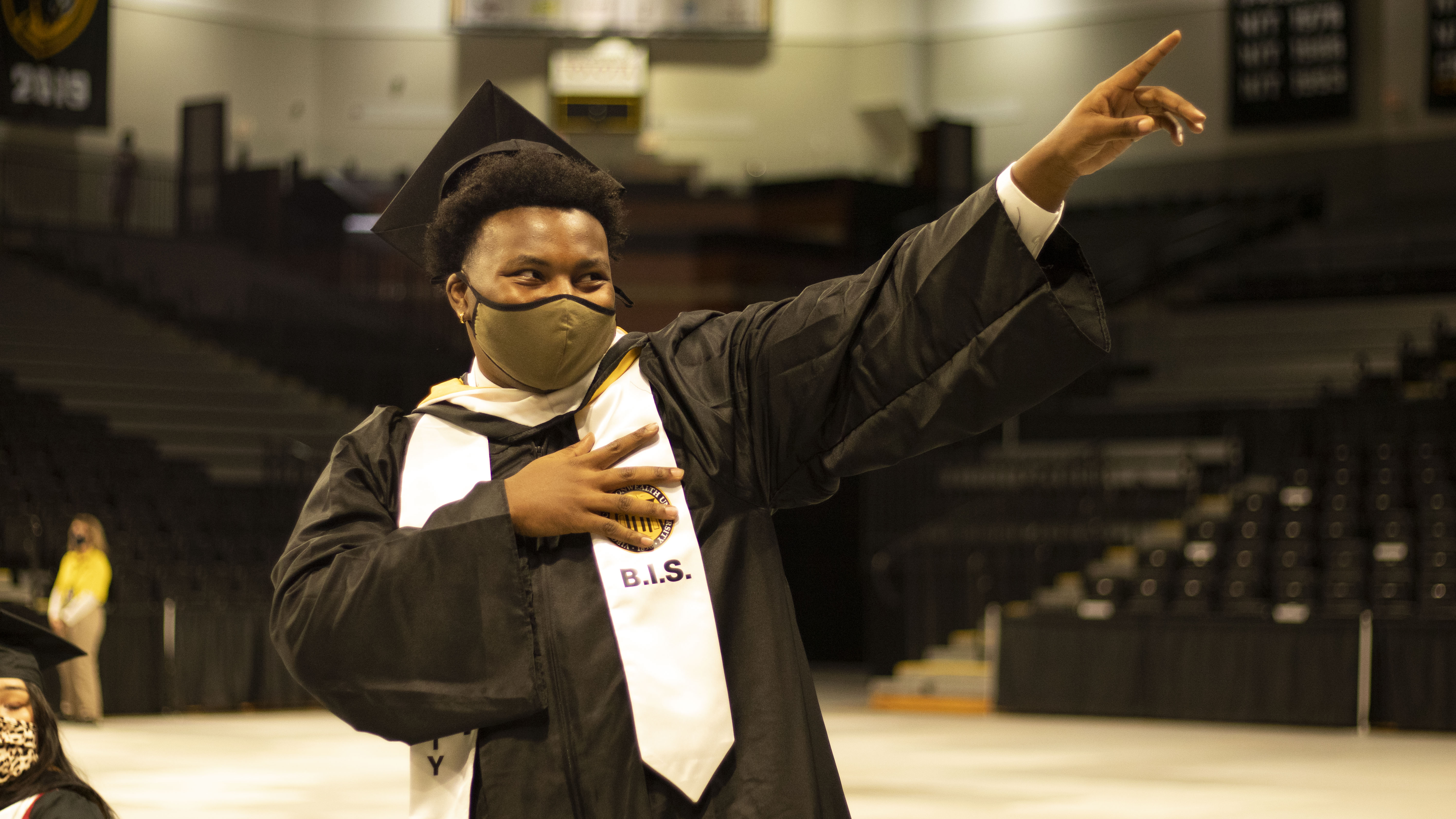 A man in graduation regalia wearing a face mask makes a hand gesture of celebration