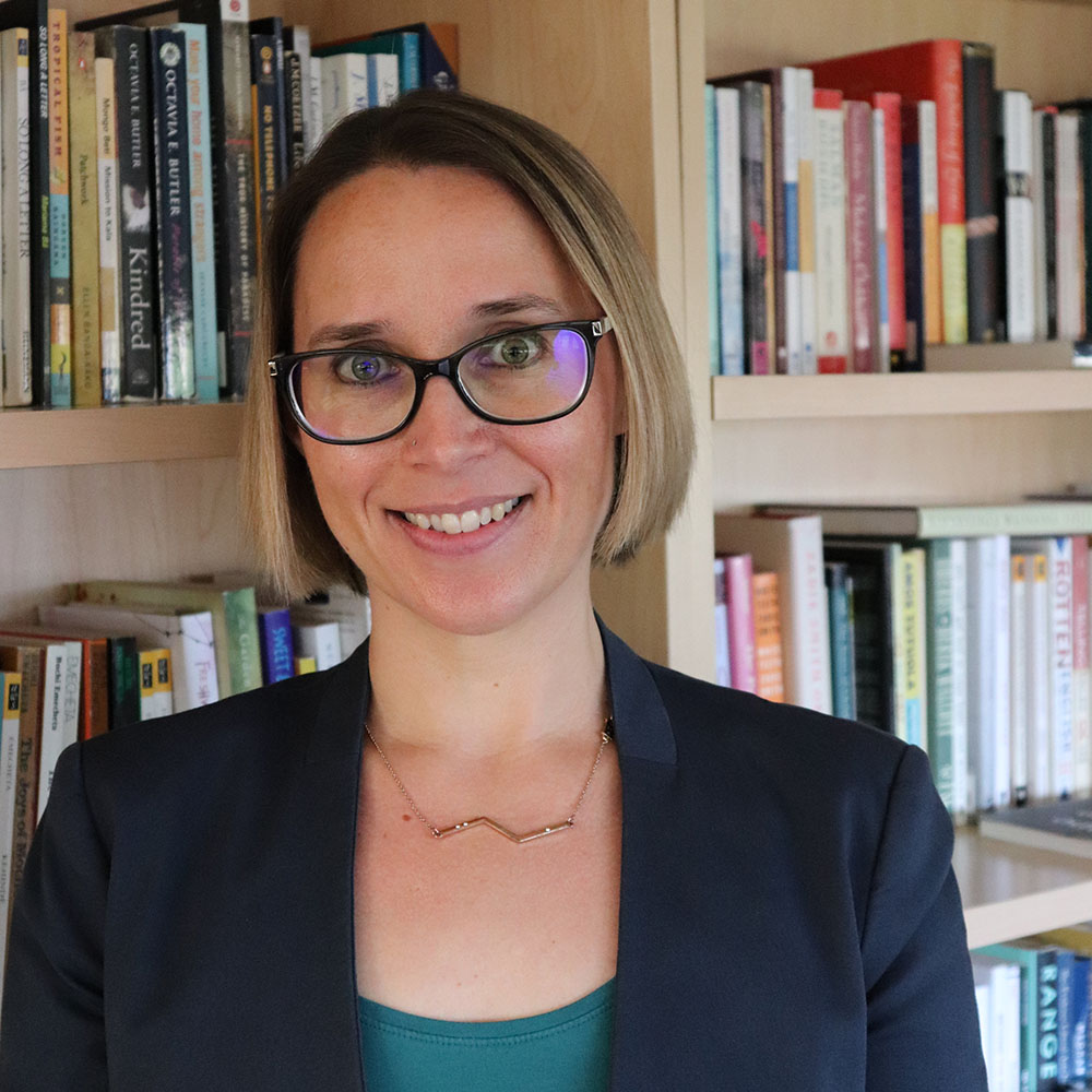 A woman wearing a teal blouse with a navy blue blazer, glasses and a gold necklace